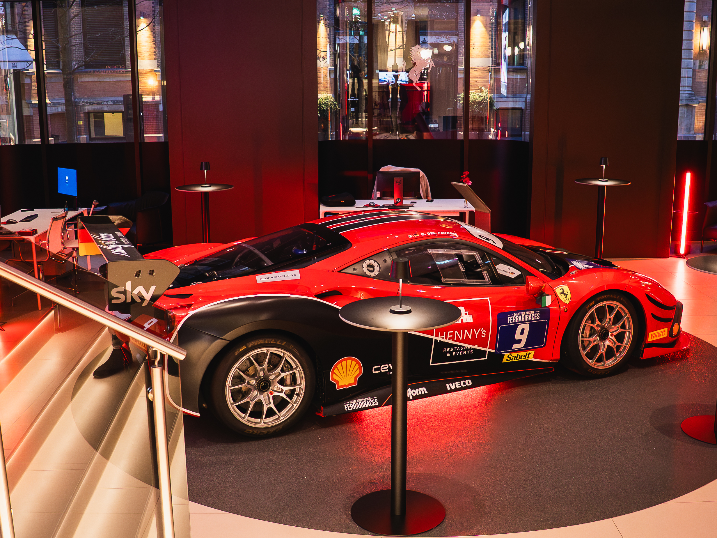 Roter Ferrari-Rennwagen in moderner Ausstellungshalle, beleuchtet von rotem Licht, mit Logos von Ferrari und Sponsoren.