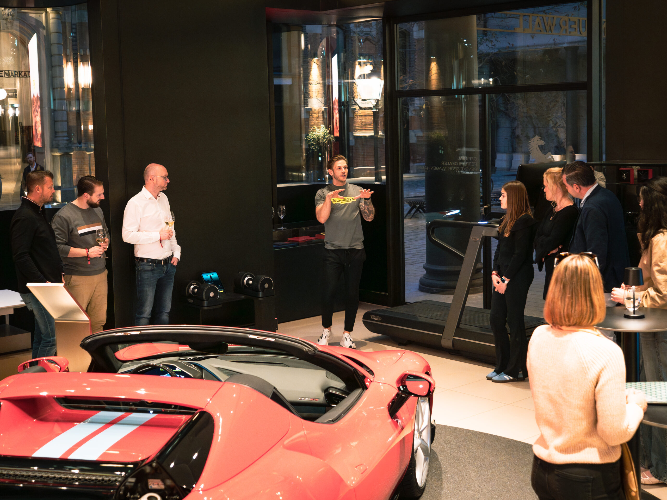 Mann hält Vortrag über Fitnessgeräte in einer luxuriösen Autolounge mit rotem Sportwagen und interessierten Zuhörern.