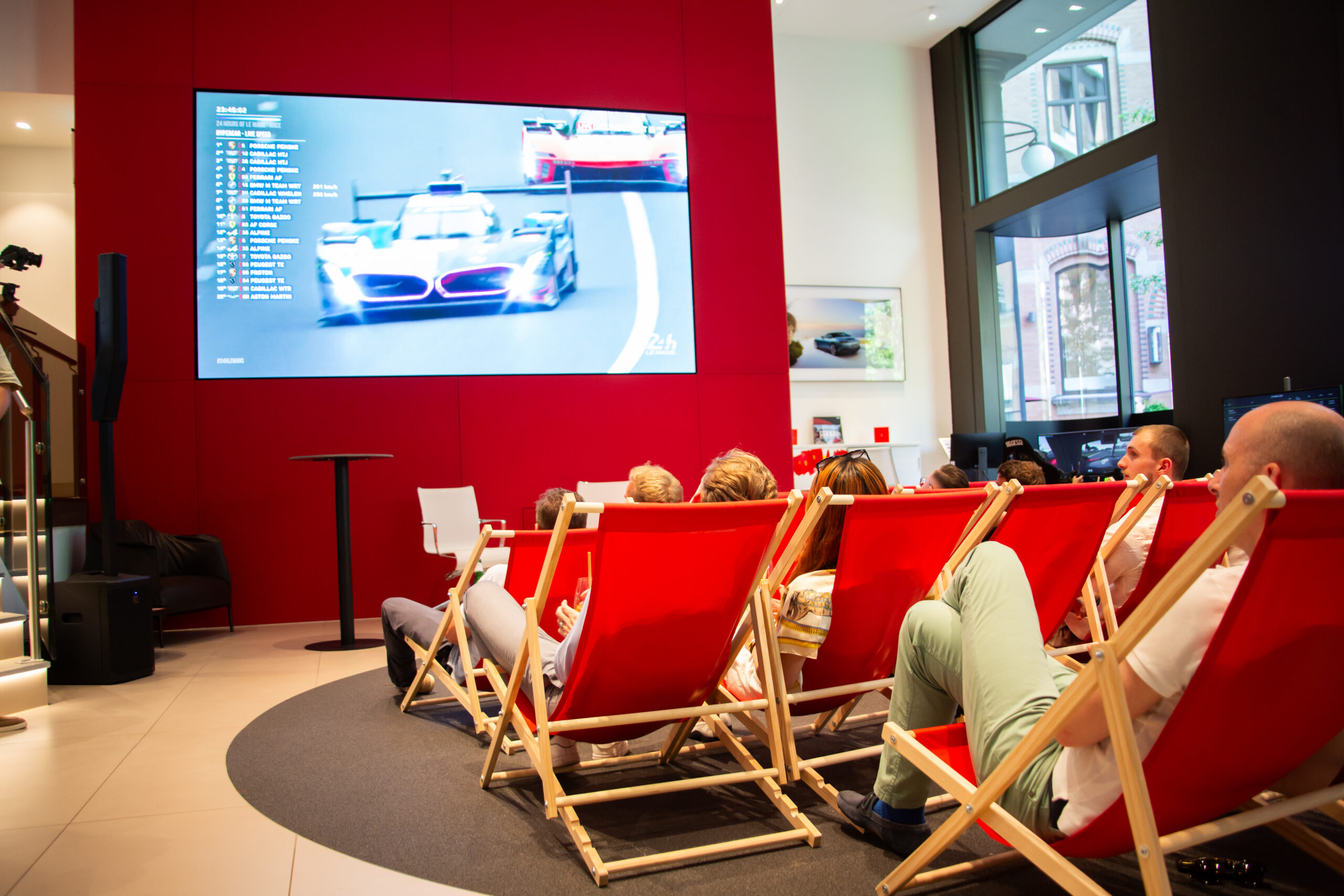 Personen schauen ein Autorennen auf einem großen Bildschirm in einem modernen, hellen Raum mit roten Liegestühlen.