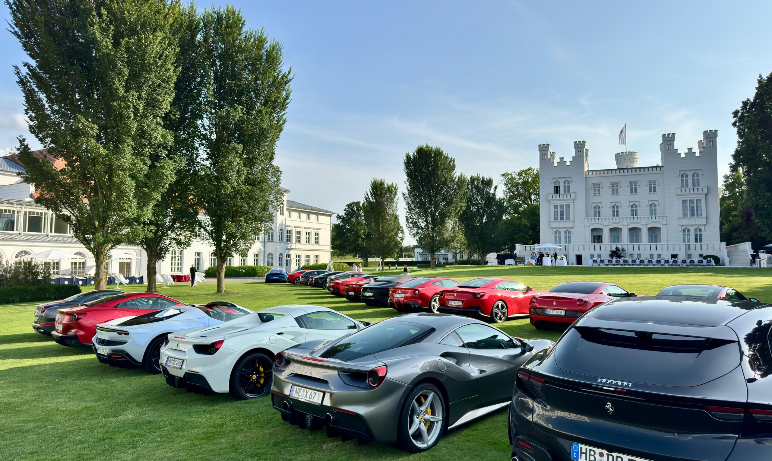 Ferraris parken vor einem weißen Schloss mit Türmen und Bäumen auf einer grünen Wiese unter blauem Himmel.