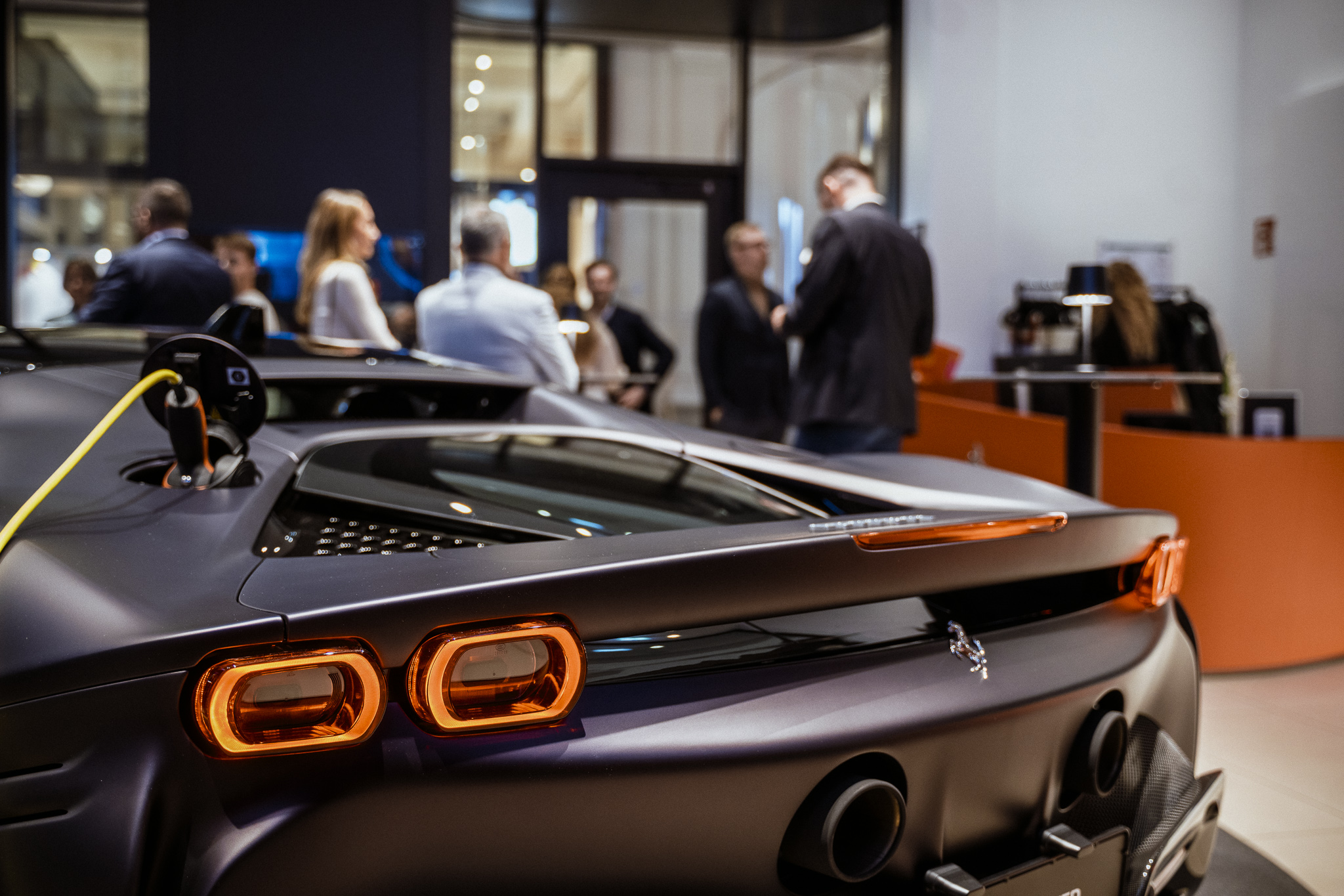 Schwarzes Elektroauto im Showroom, Publikum im Hintergrund. Moderne Fahrzeugpräsentation mit Ladeanschluss sichtbar.