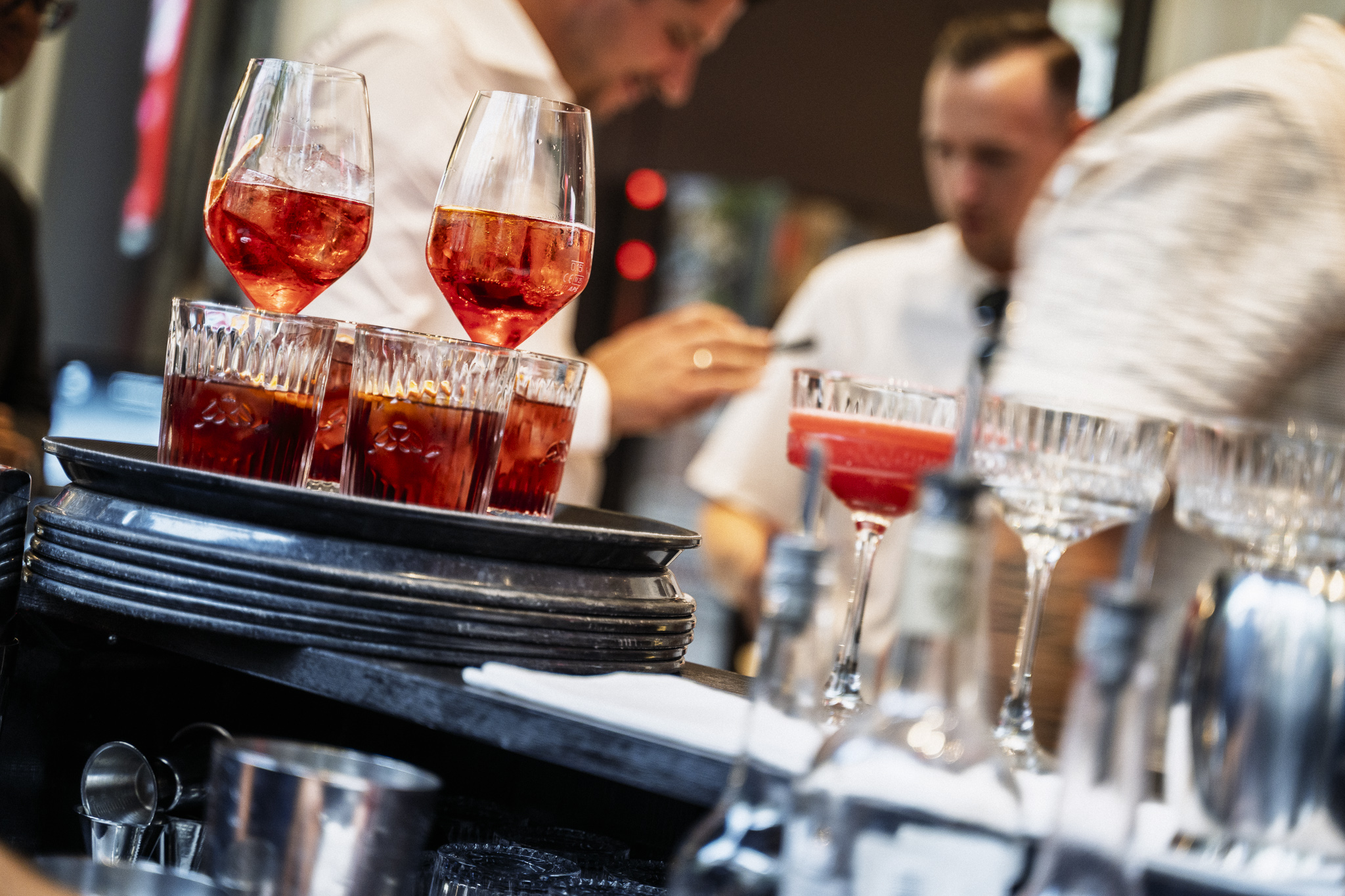 Rote Cocktails in eleganten Gläsern auf einem Tablett in einer Bar, mit unscharfen Barkeepern im Hintergrund.