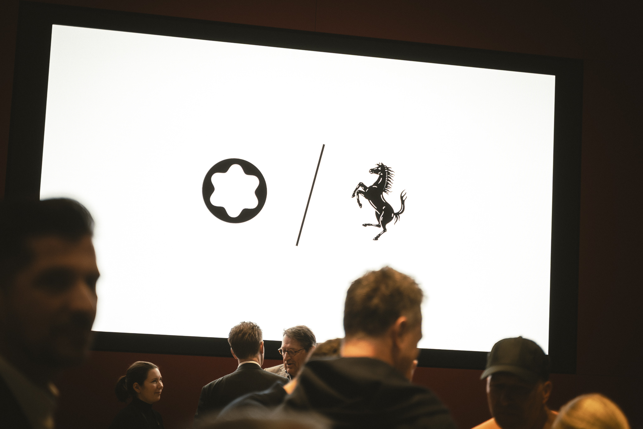 Menschenmenge vor einem Bildschirm mit Montblanc und Ferrari Logos, zeigt eine Partnerschaft oder Veranstaltung.