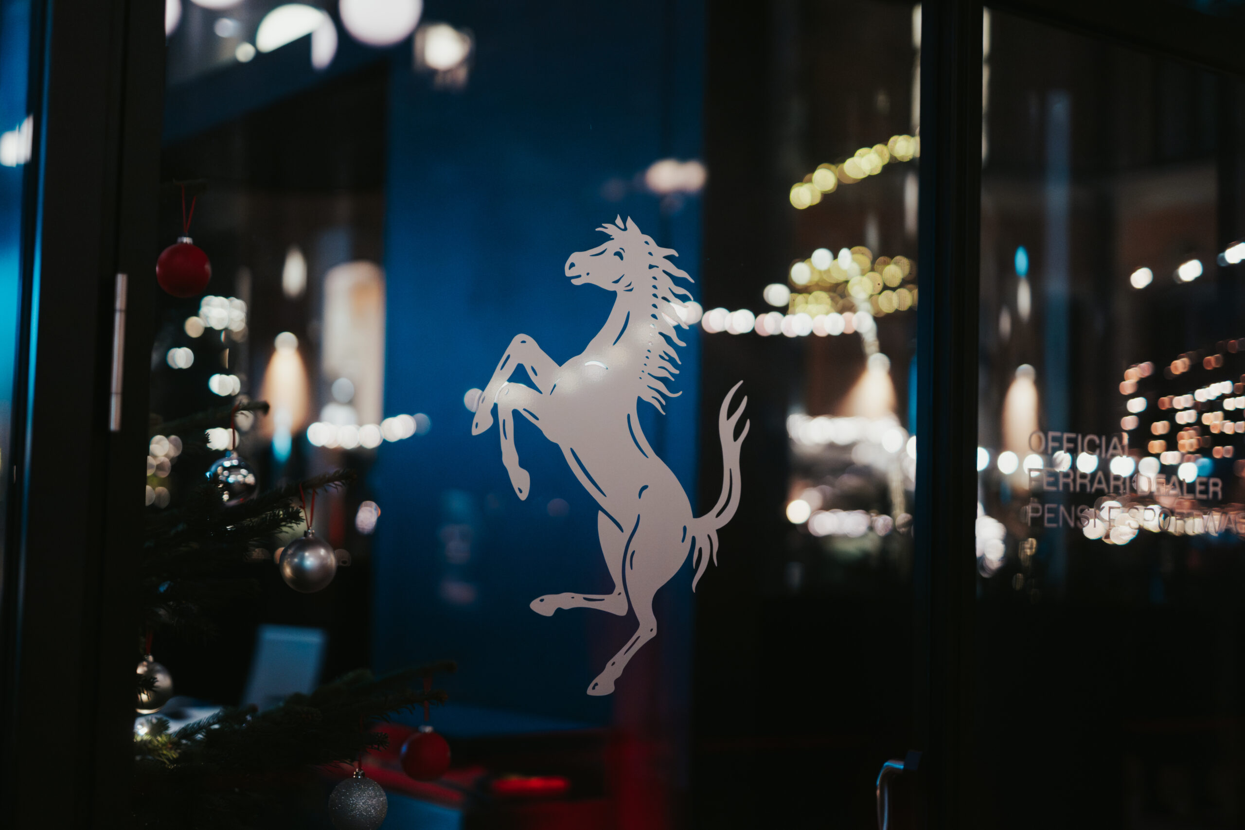 Ferrari-Logo auf einer Glasoberfläche bei Nacht mit Weihnachtsdekoration im Hintergrund.