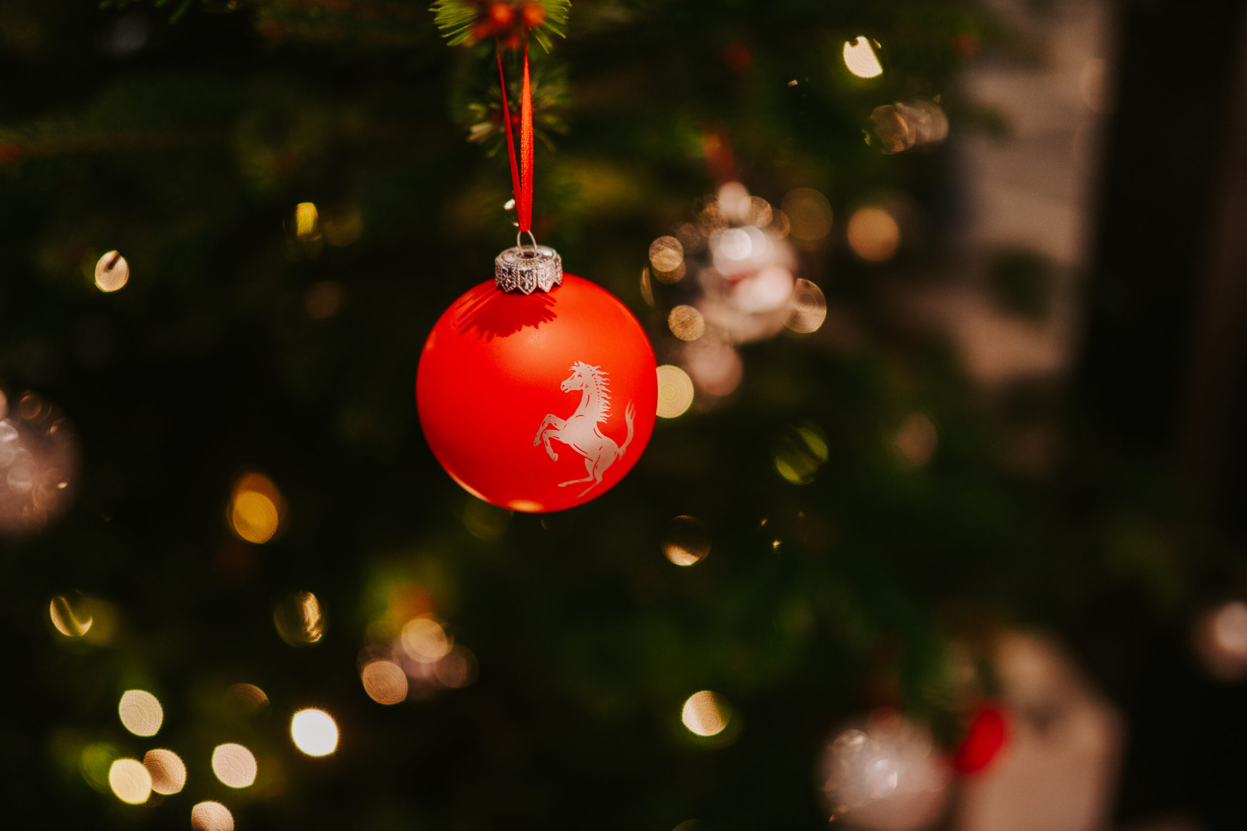 Rote Weihnachtskugel mit springendem Pferd-Logo an geschmücktem Weihnachtsbaum mit Lichtern im Hintergrund.