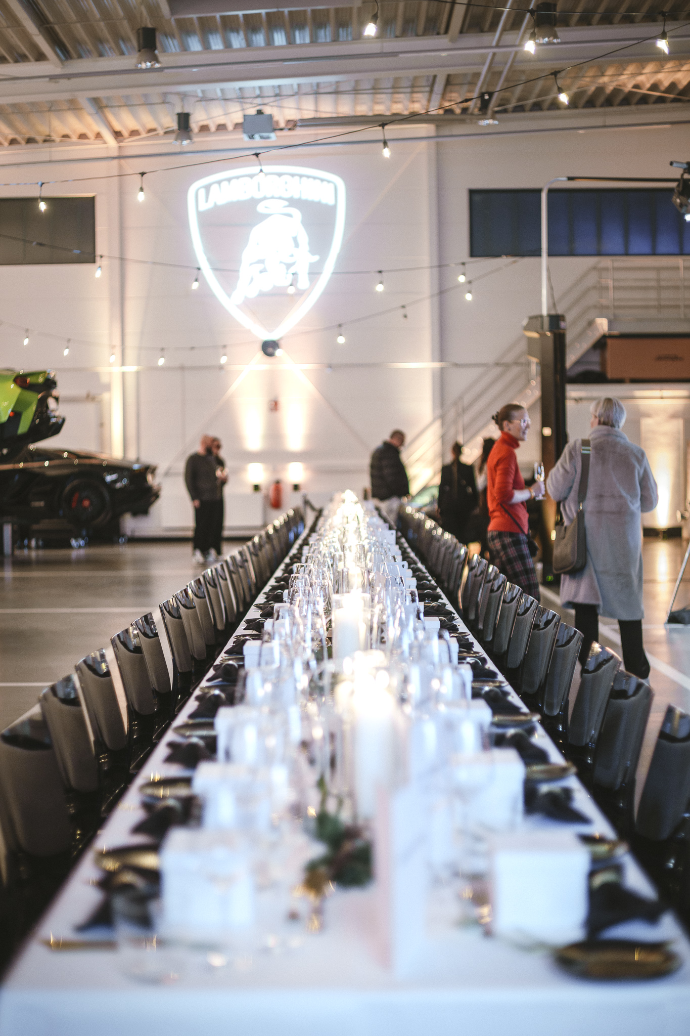 Elegant gedeckter langer Tisch in einer Halle mit Lamborghini-Logo an der Wand, Personen im Hintergrund.