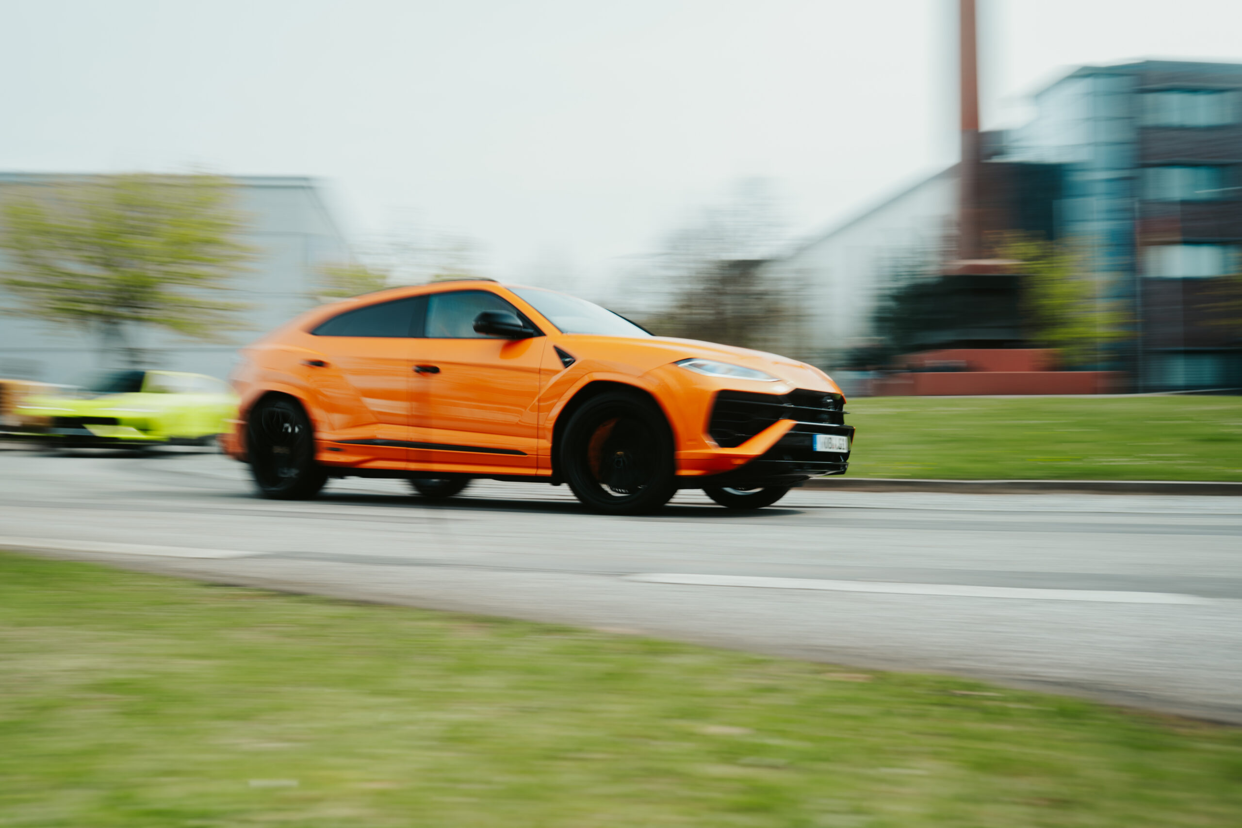 Orangefarbener Lamborghini Urus fährt schnell auf der Straße, unscharfe Gebäude im Hintergrund.